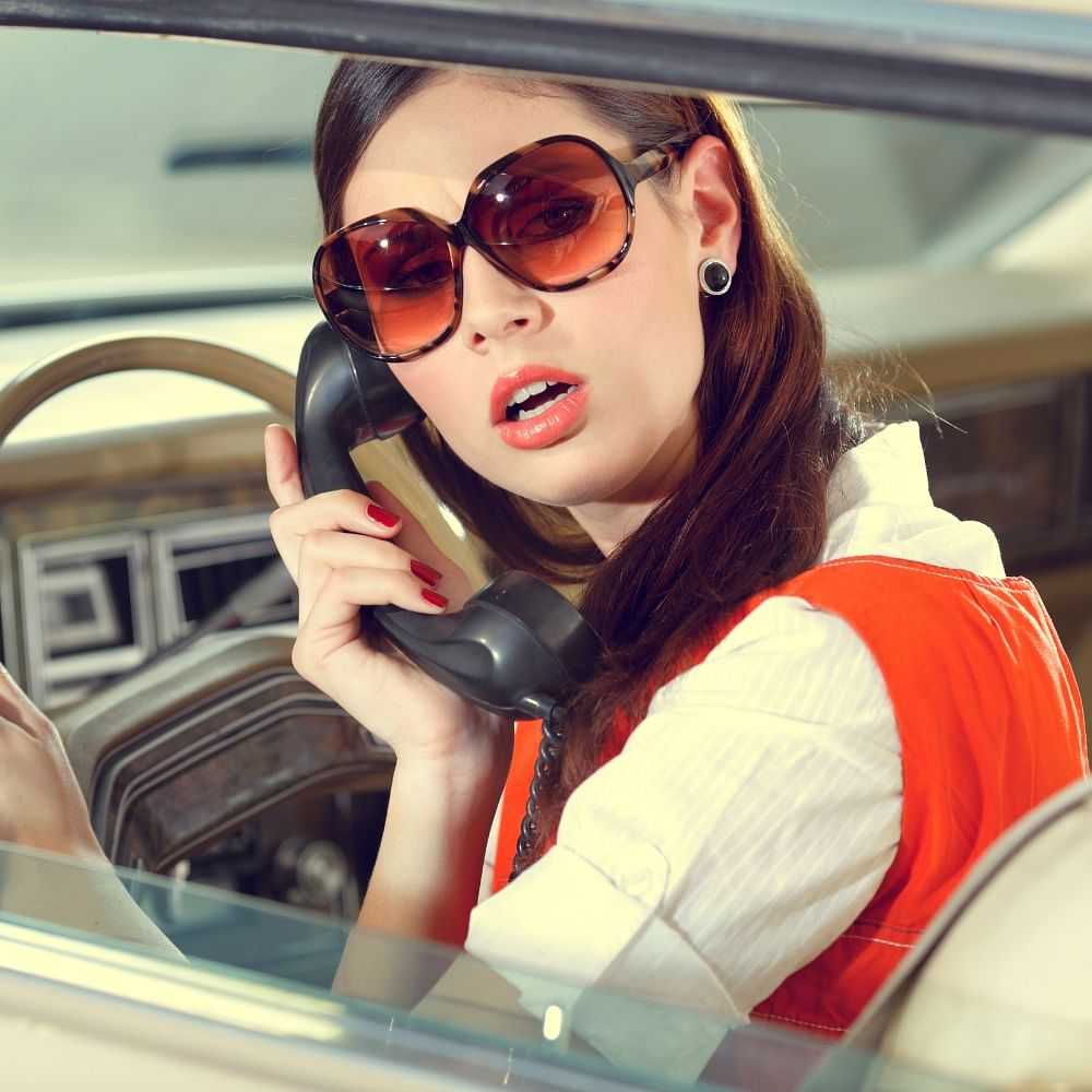 Woman in sunglasses talking on a vintage car phone, looking surprised.